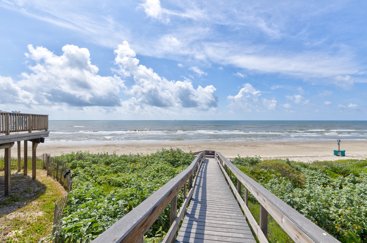 A wooden boardwalk leads through coastal greenery to a pristine sandy beach, offering direct access to the waves and shoreline under open blue skies.