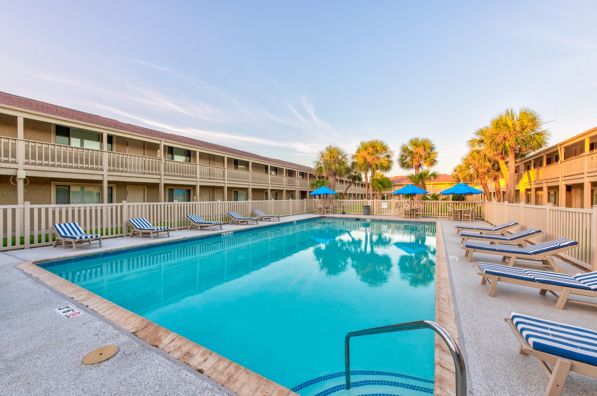 Two-story property buildings surround a crystal-clear swimming pool with palm trees and colorful umbrellas creating a tropical oasis.
