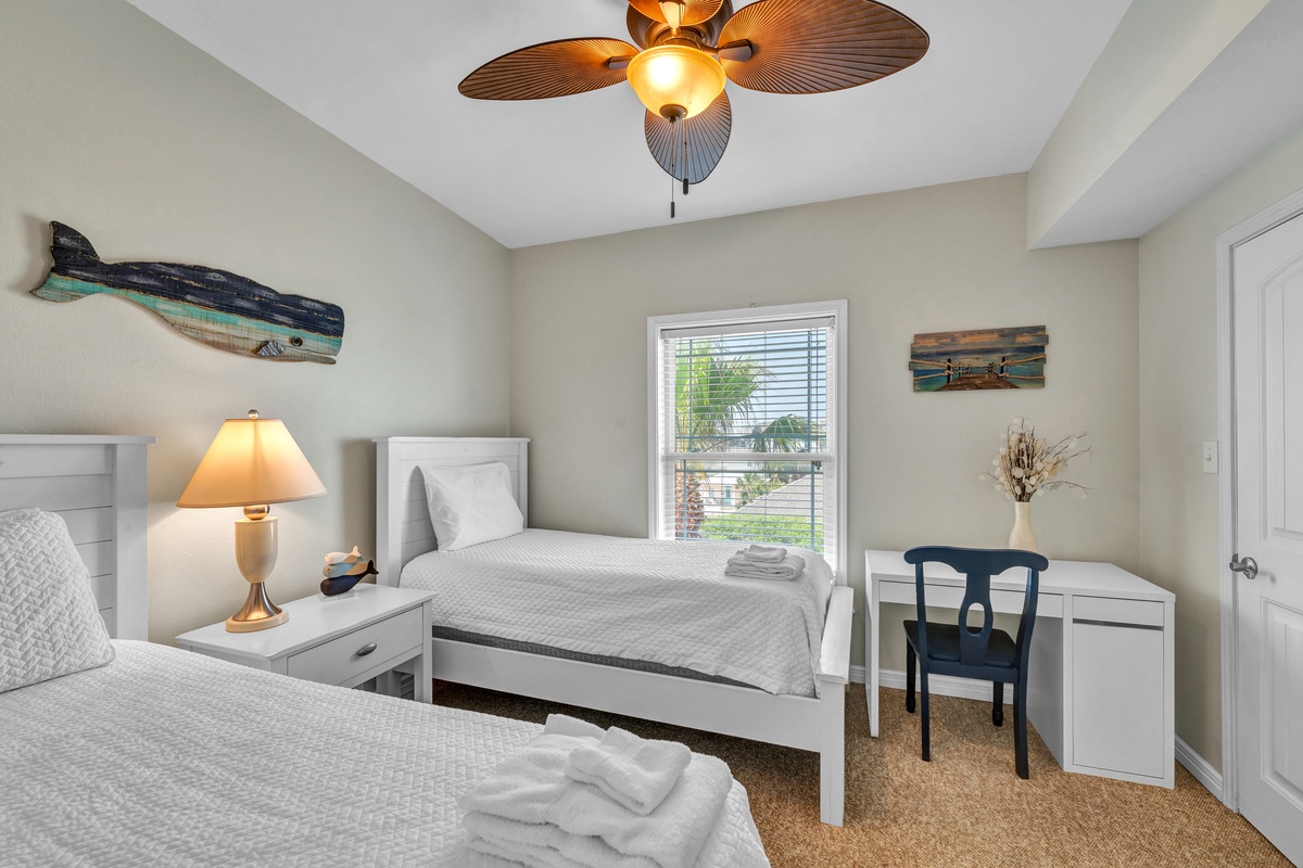 Your coastal retreat features twin beds with crisp linens, beach-themed artwork, and a cozy workspace beneath the tropical ceiling fan.