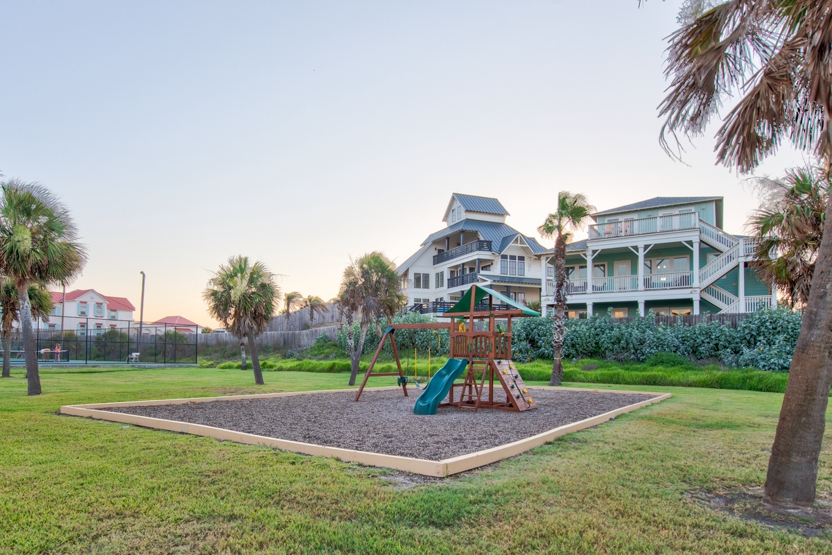 Coastal vacation homes surround a dedicated playground area with palm trees and recreational facilities nearby.