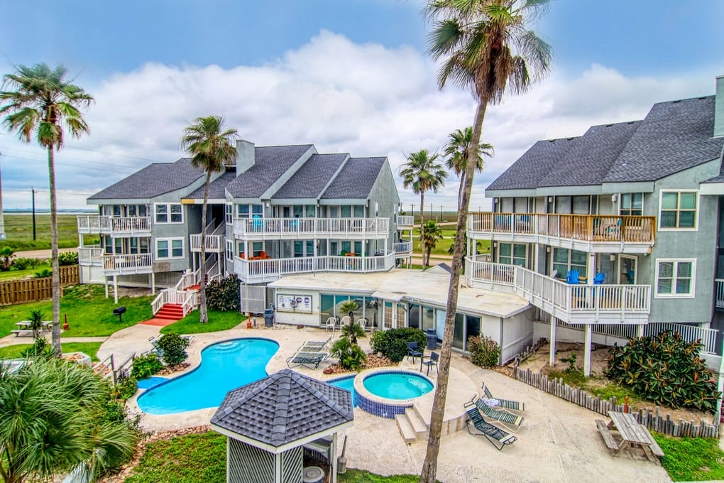 Coastal resort complex featuring multiple buildings with balconies, swimming pool, and palm trees beneath blue skies.
