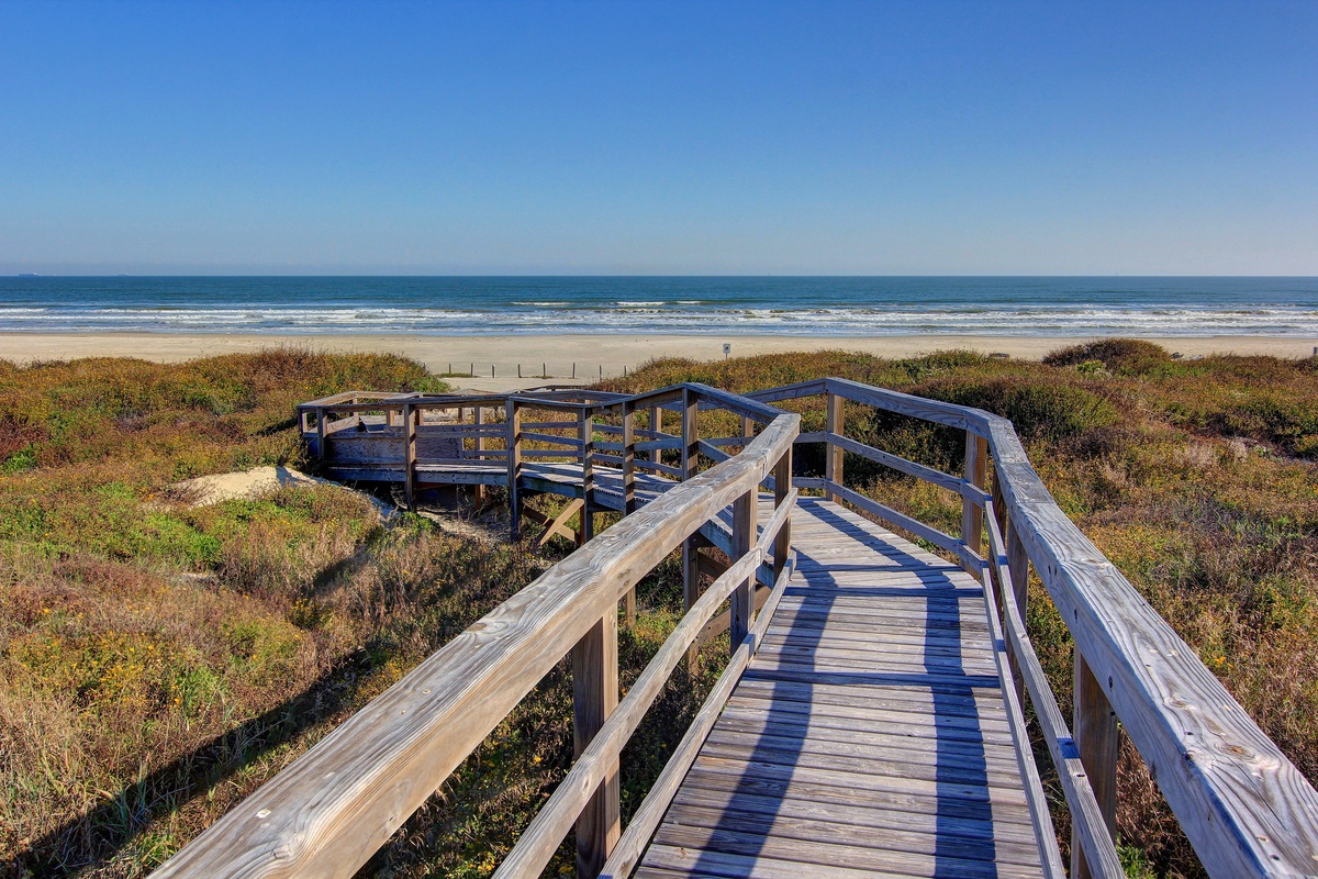 A wooden boardwalk leads to pristine sandy beach and ocean waves under clear blue skies.