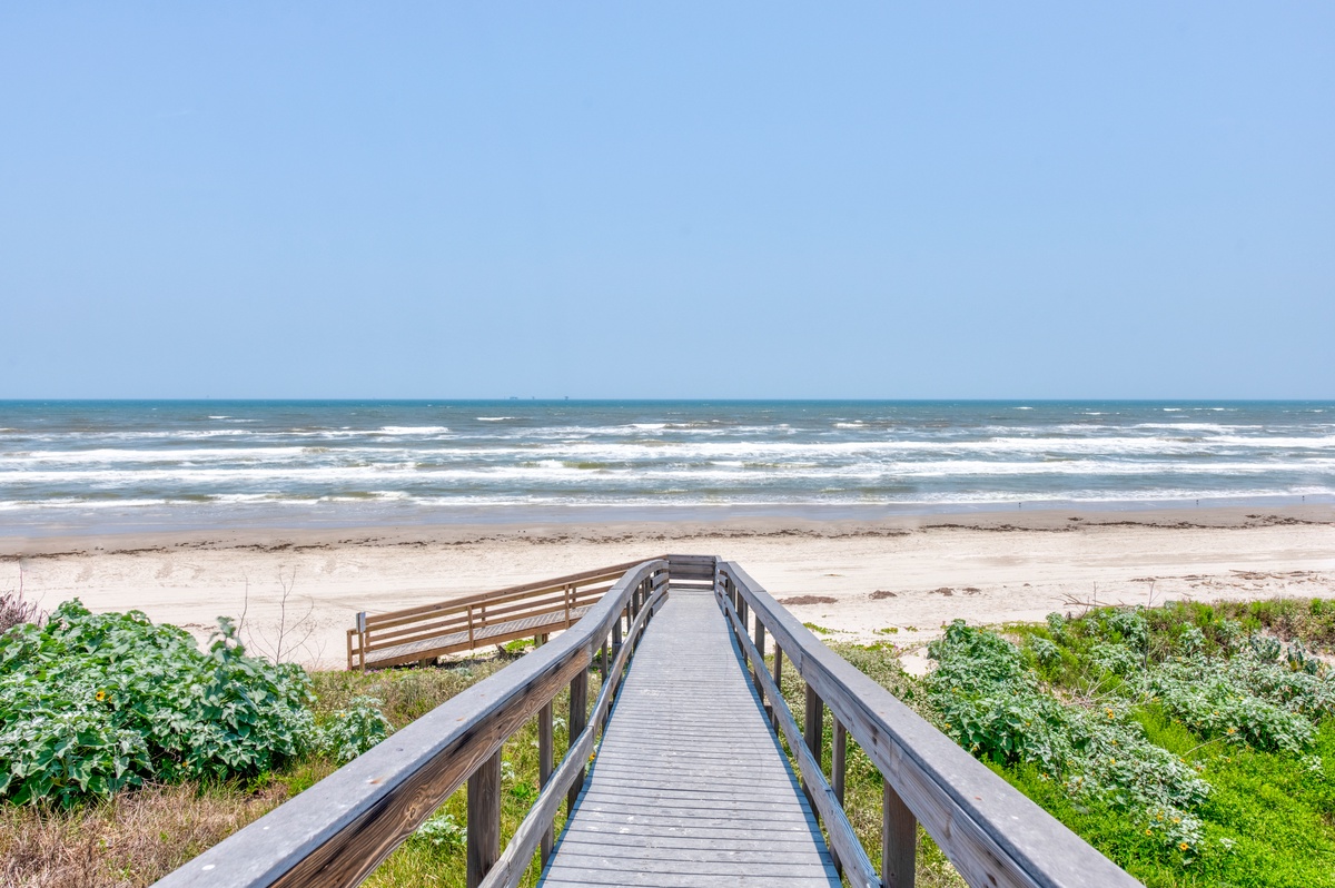 A wooden boardwalk leads to a pristine sandy beach with gentle waves rolling onto shore under a clear blue sky—ideal coastal escape nearby.