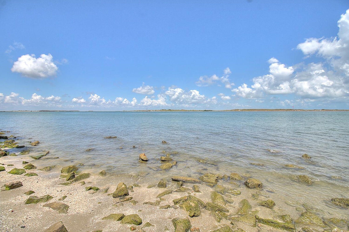 Crystal clear waters meet pristine shoreline dotted with natural rocks under expansive blue skies and puffy white clouds.