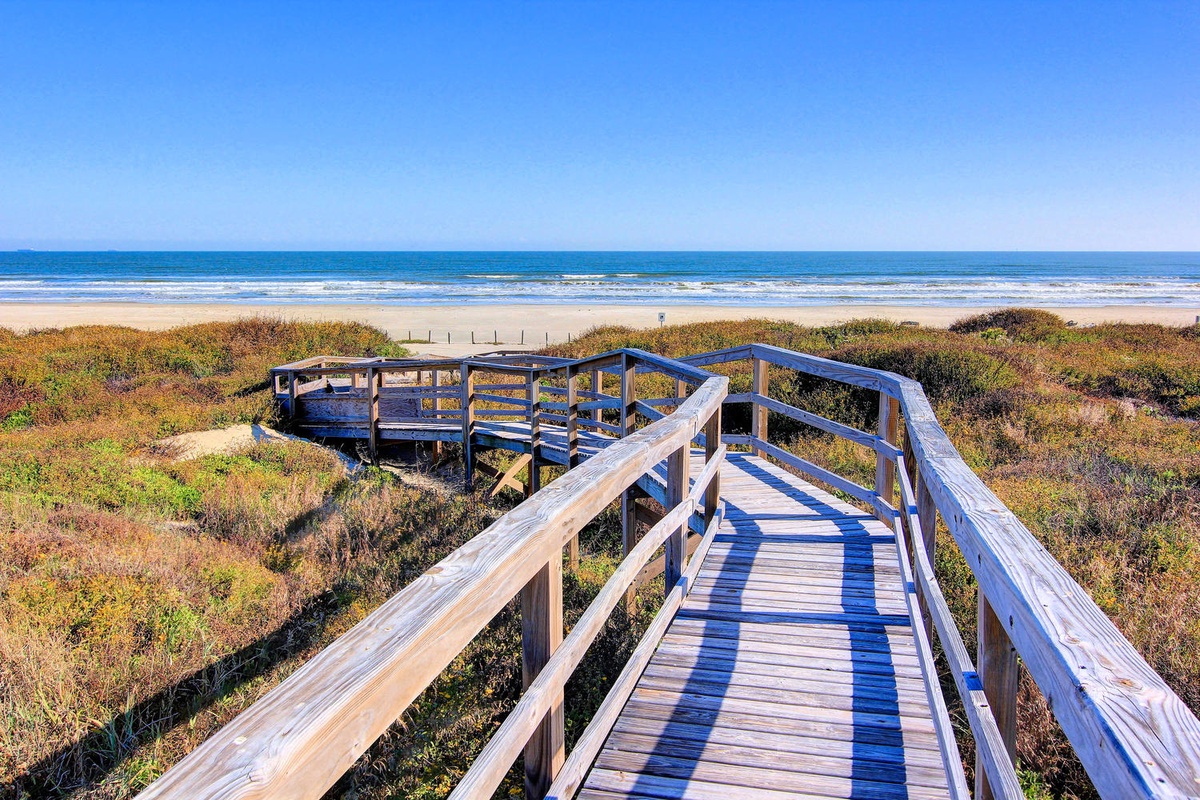 Wooden boardwalk winds through coastal dunes, leading to pristine sandy beach and sparkling blue ocean waters.