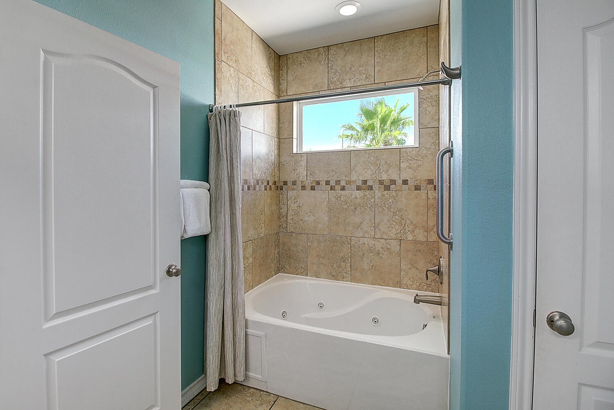 Unwind in your private spa-like bathroom featuring a luxurious jetted tub surrounded by elegant stone tile, with tropical views through the window.