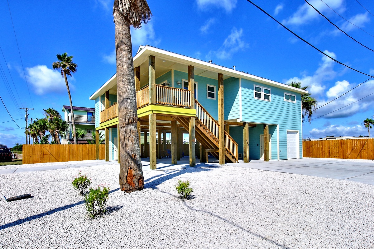 Bright turquoise beach house on stilts with tropical palms and white sand surroundings in a coastal neighborhood.