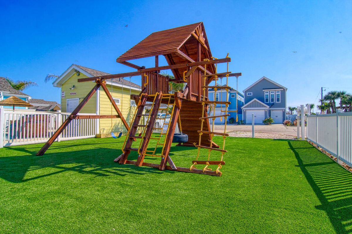 A stunning playground structure on pristine artificial turf, nestled in a vibrant coastal community with charming beach houses.