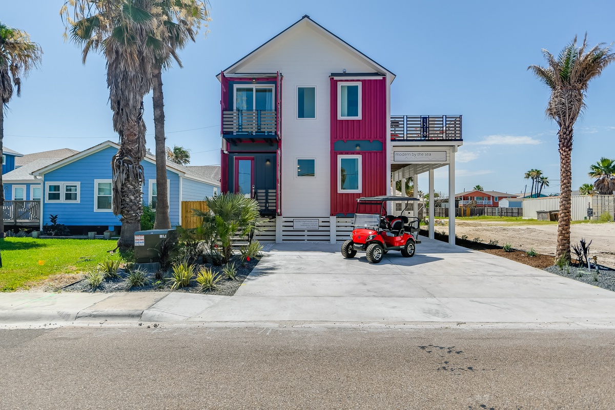 Modern beachside property featuring distinctive red and white architecture with covered parking and tropical landscaping in a coastal neighborhood setting.