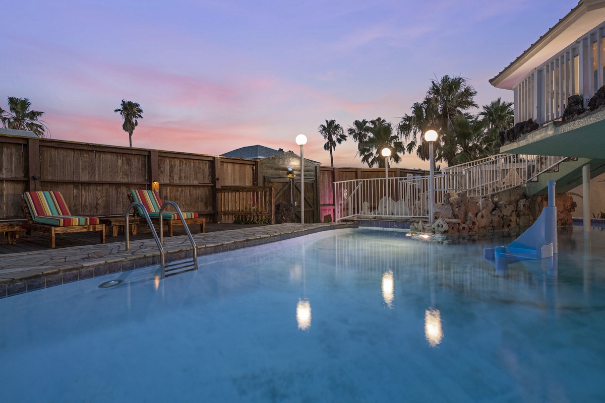Tropical pool area with colorful lounge chairs under a stunning pink sunset sky, surrounded by palm trees and vacation vibes.