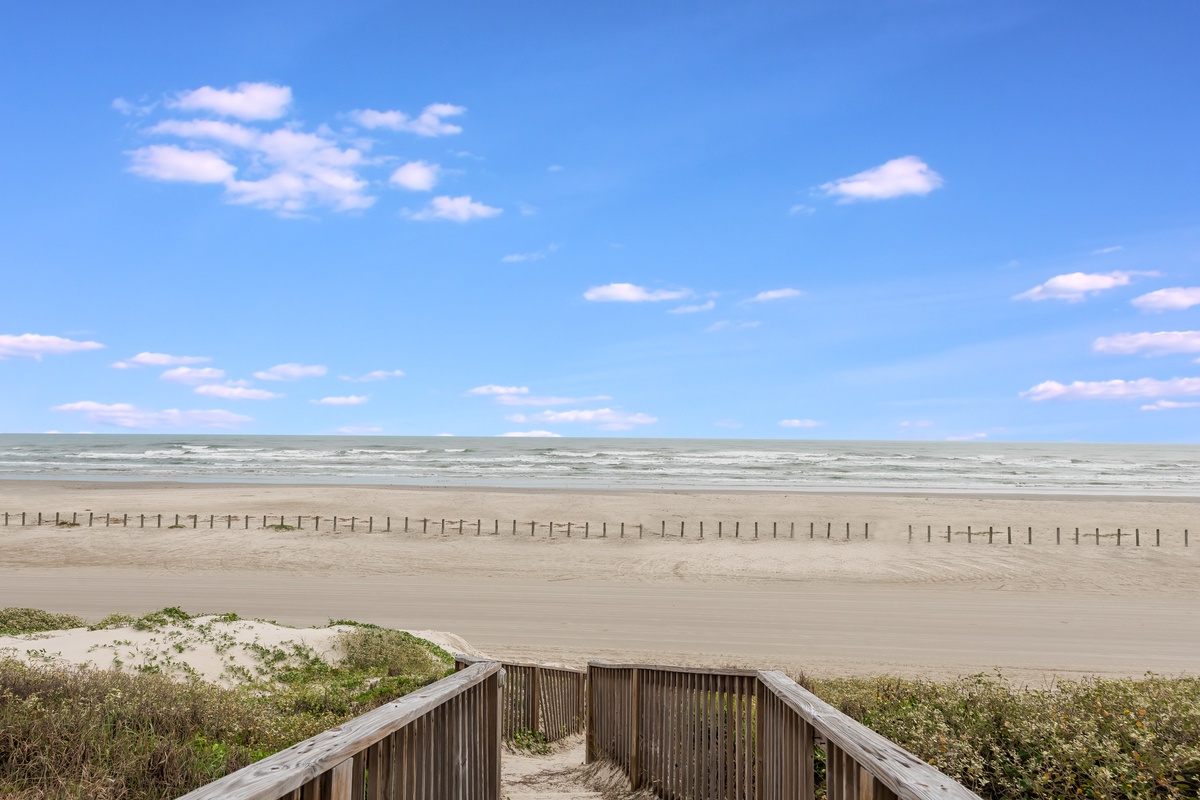 Wooden boardwalk leads directly to pristine sandy beach with gentle waves and endless blue sky dotted with fluffy white clouds.