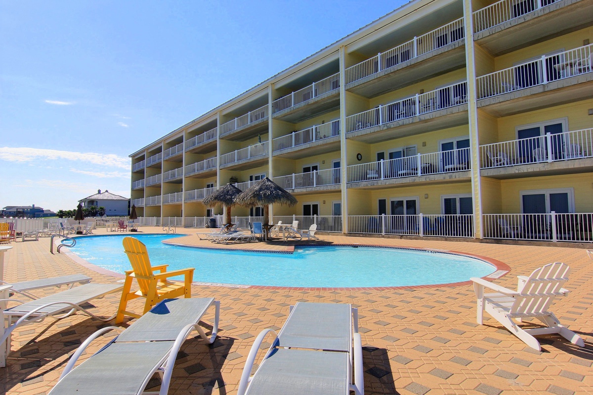 Modern resort building surrounds an inviting pool area with comfortable loungers and tropical tiki bar, creating a perfect vacation atmosphere.