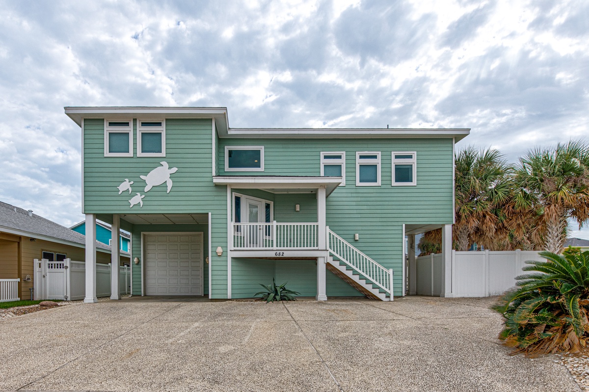 Modern coastal home featuring sea turtle artwork, elevated design with covered parking, and tropical landscaping in a beach community setting.