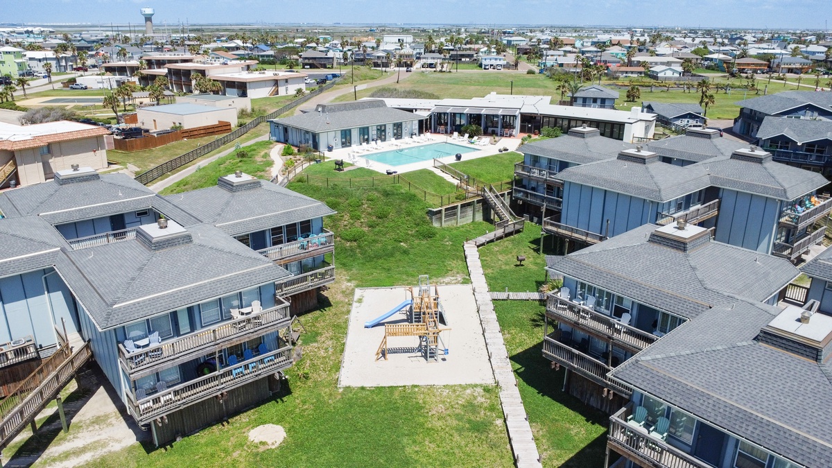 Aerial view of coastal vacation rental community featuring beach houses, swimming pool, and playground facilities in a residential neighborhood setting.