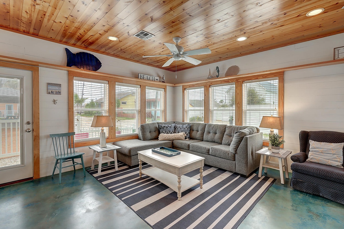 Relax in your coastal-inspired living room with warm wood ceilings, comfy sectional seating, and bright windows flooding the space with natural light.