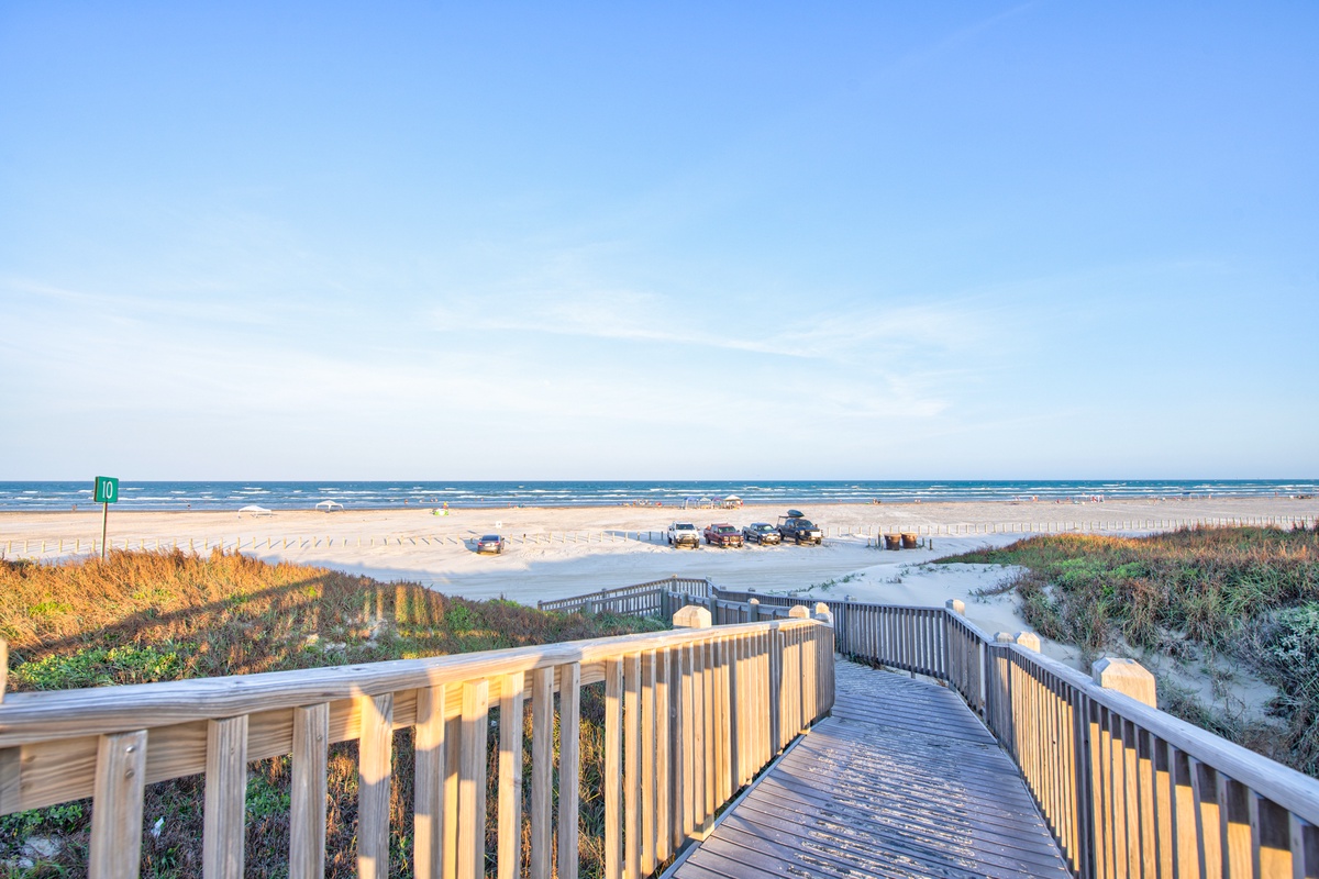Wooden boardwalk leads to pristine sandy beach with crystal-clear ocean waters under endless blue skies.