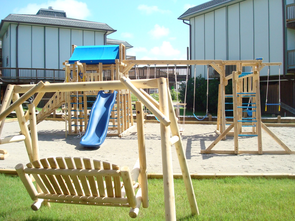Expansive wooden playground with slides, swings, and climbing features set against modern vacation rental buildings.