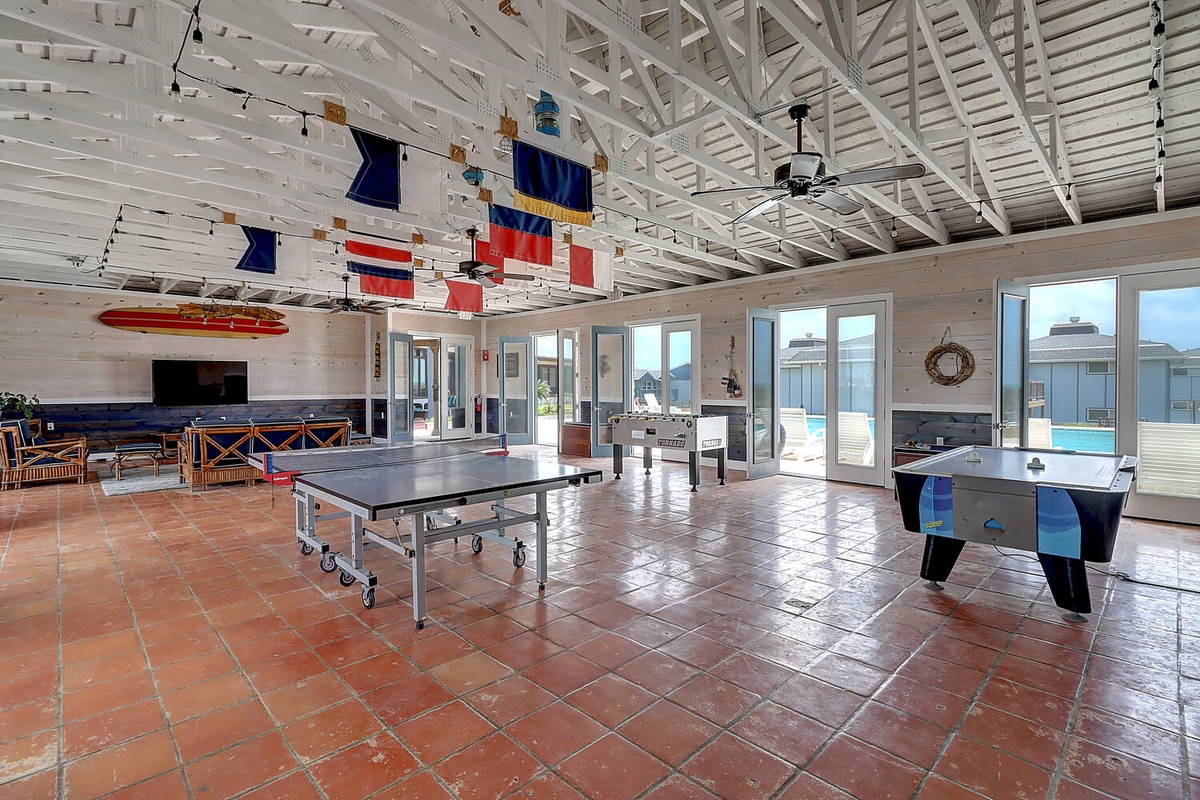 Your game room awaits with ping pong, air hockey, and nautical charm beneath soaring white beams and colorful maritime flags.