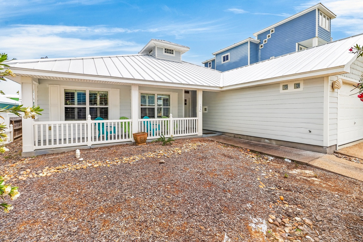 Charming coastal home with white railings and metal roof, nestled among blue-trimmed houses in a peaceful beach neighborhood setting.
