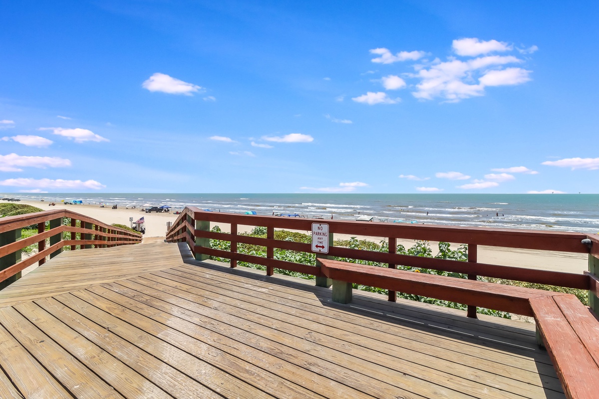 Wooden boardwalk overlooks sandy beach with ocean waves and scattered beachgoers under a bright blue sky.