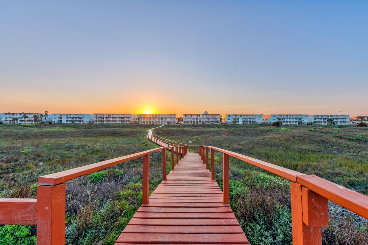 A scenic wooden boardwalk winds through coastal marshland toward beachfront condominiums, bathed in golden sunset light.
