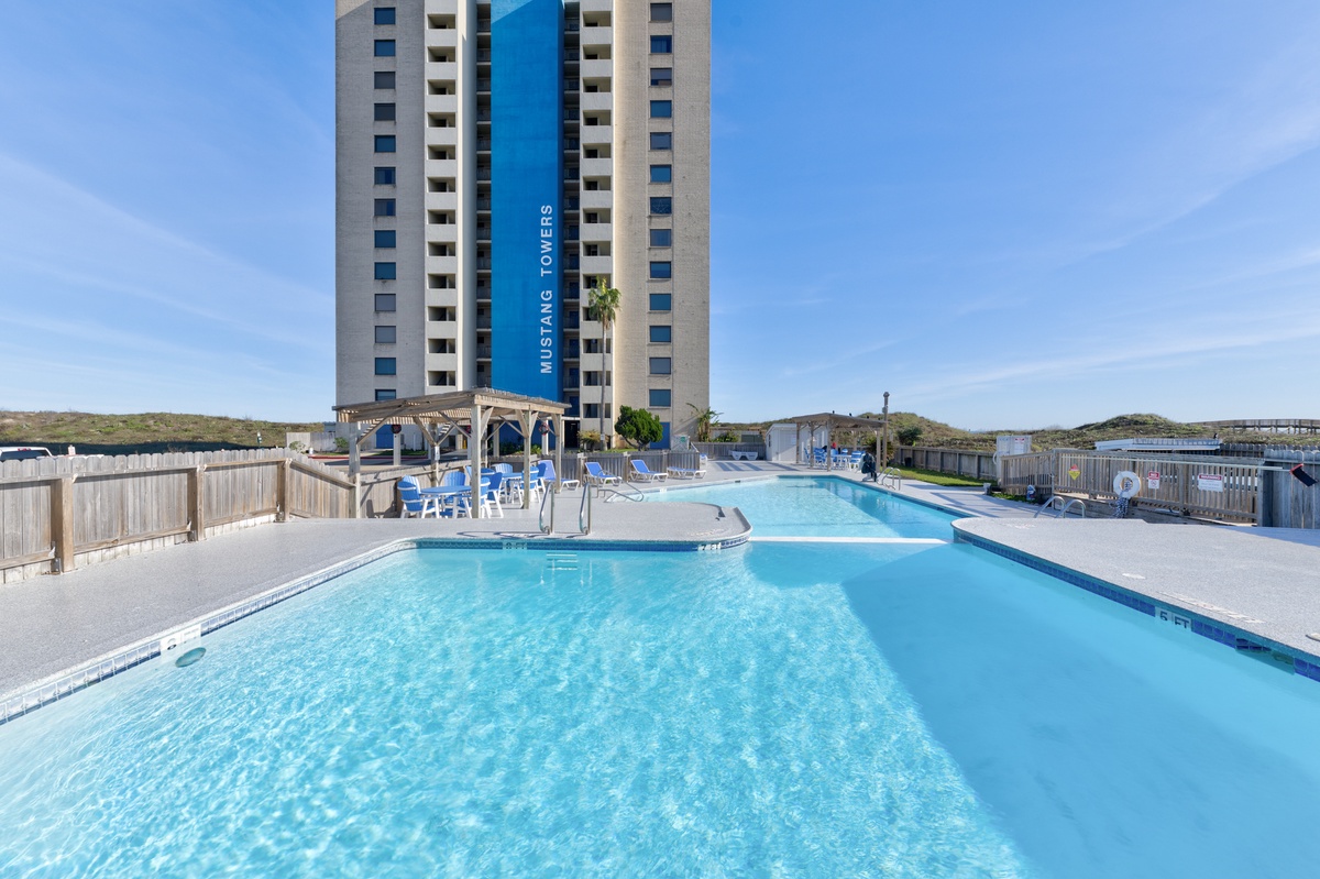 Large outdoor pool area with clear blue water and seating, set against the backdrop of Mustang Towers resort building.