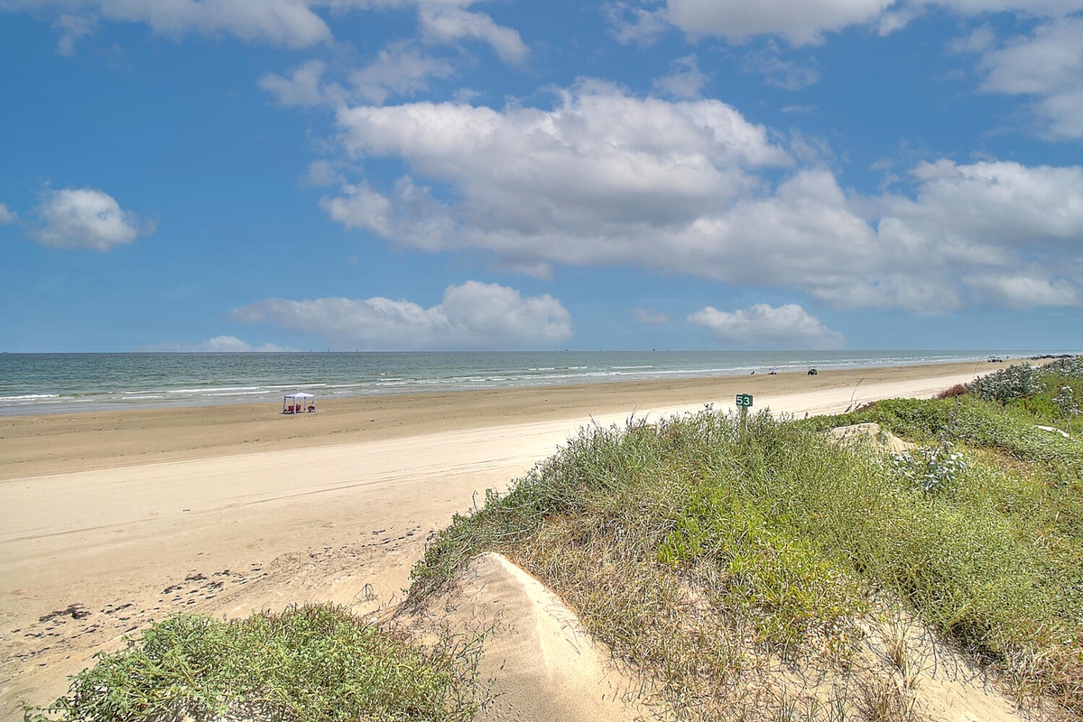 Pristine sandy beach stretches along the coastline with natural dunes and vegetation under a brilliant blue sky with scattered clouds.