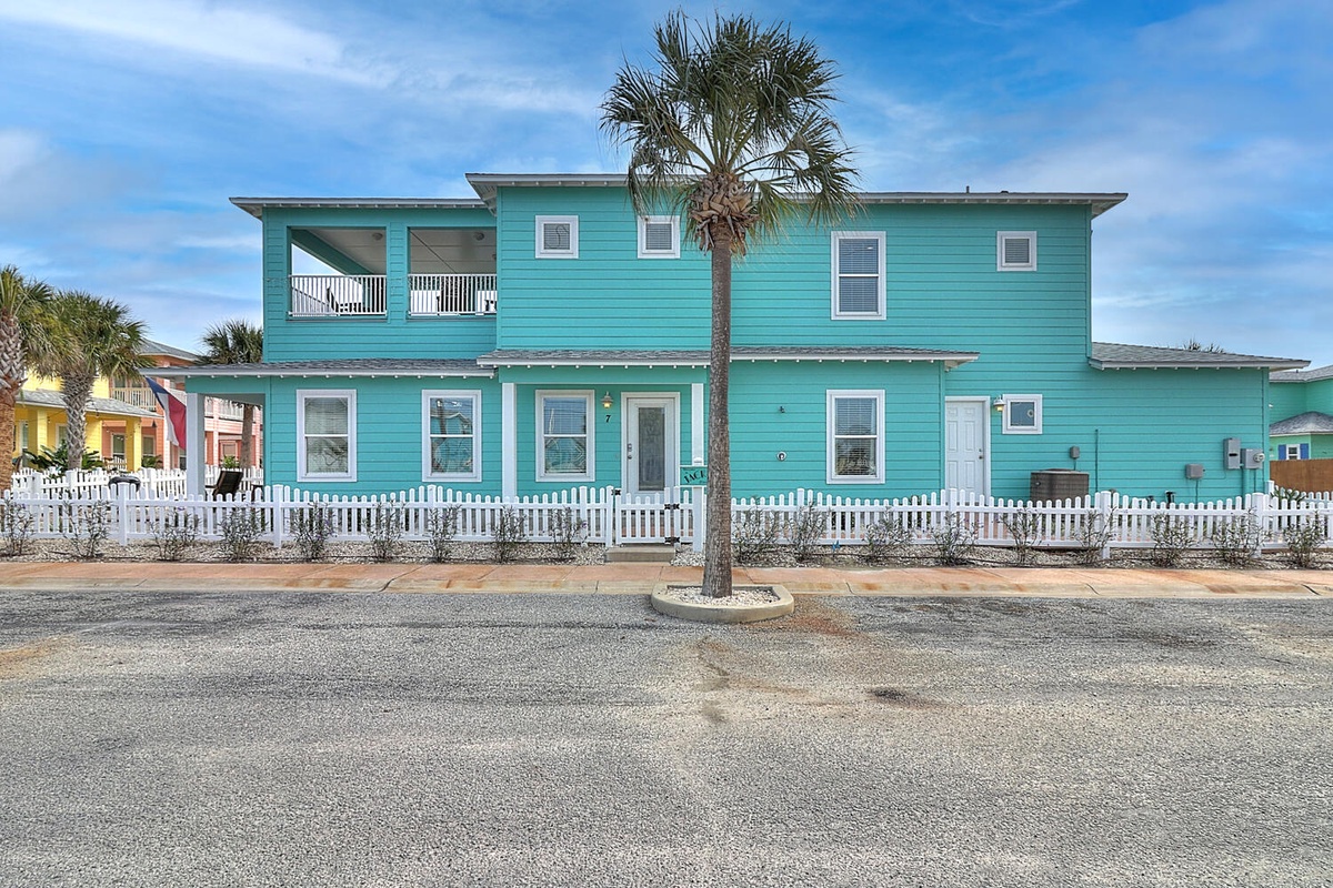 Bright turquoise beach house with white picket fence, palm tree, and colorful neighborhood charm creates the perfect coastal getaway.