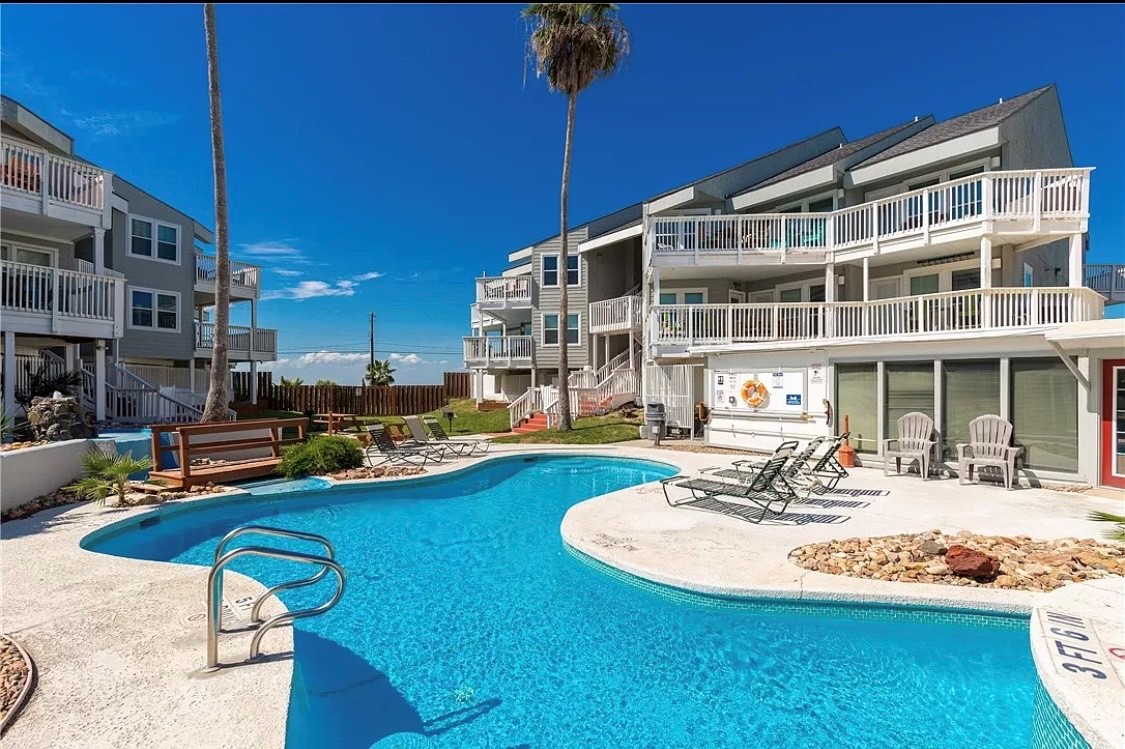 Beachfront resort featuring a curved swimming pool with multiple buildings, balconies, and tropical landscaping under clear blue skies.