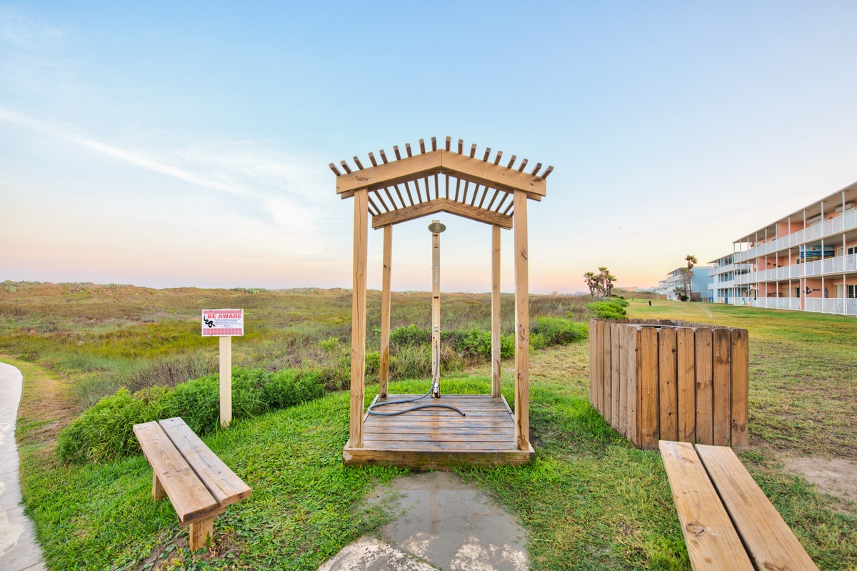 A wooden shelter structure sits in natural grassland with modern apartment buildings visible in the distance.