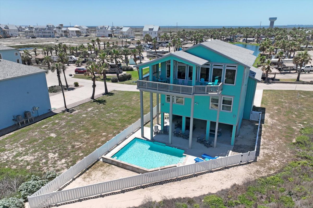 Elevated turquoise beach house with private pool, surrounded by palm trees and coastal community near the shore.