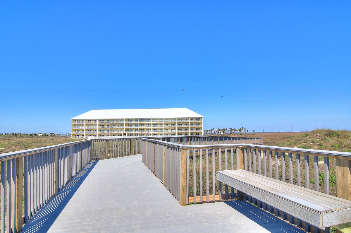 Elevated boardwalk leads to a beachfront hotel complex, offering scenic views across coastal dunes and natural vegetation under clear blue skies.