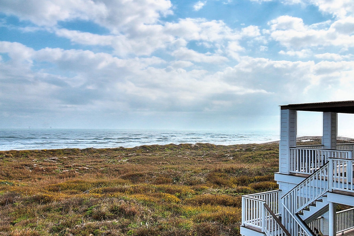 Coastal property features elevated deck overlooking natural dunes and ocean waves under dramatic cloudy skies.