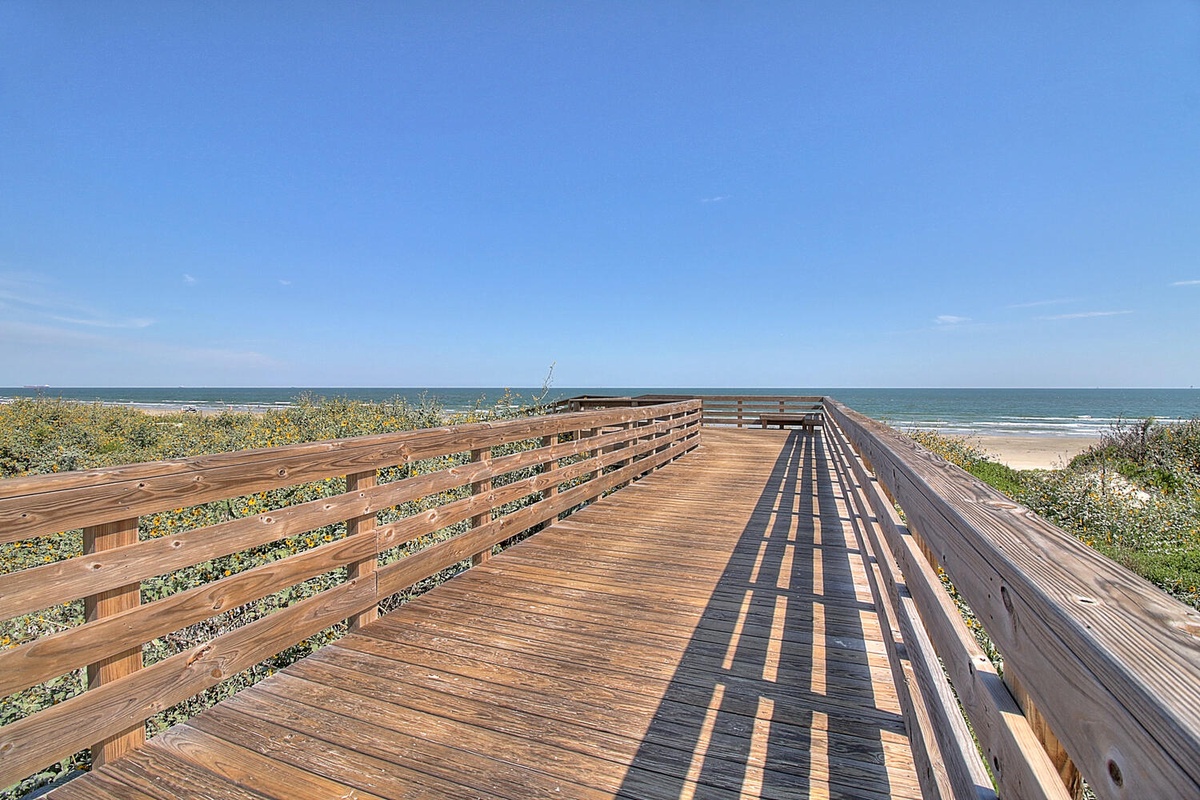 Wooden boardwalk leads through coastal dunes to pristine beach under clear blue skies.
