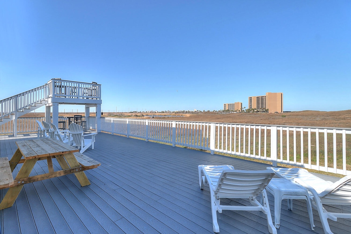 Elevated deck with white lounge chairs and picnic table overlooking coastal landscape and distant buildings under clear blue sky.
