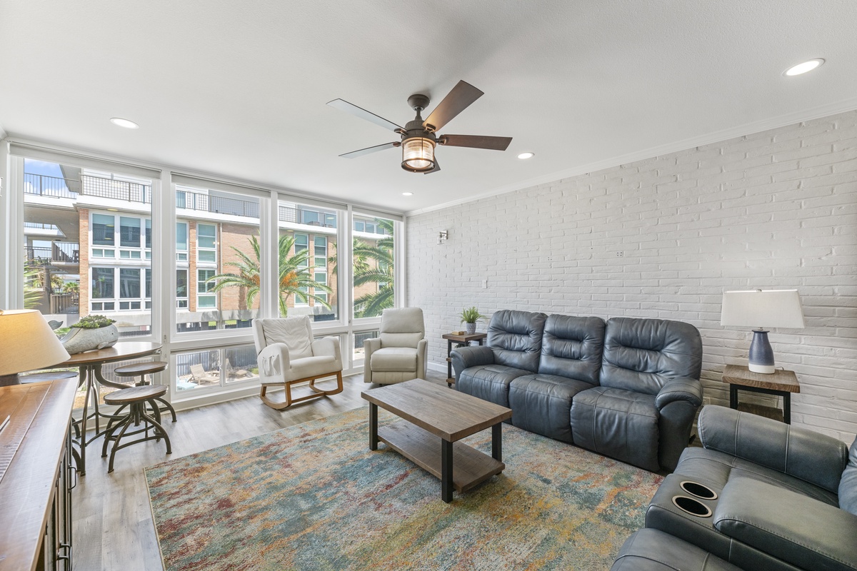 Relax in your spacious living room featuring plush leather seating, stylish brick accent wall, and bright city views through expansive windows.