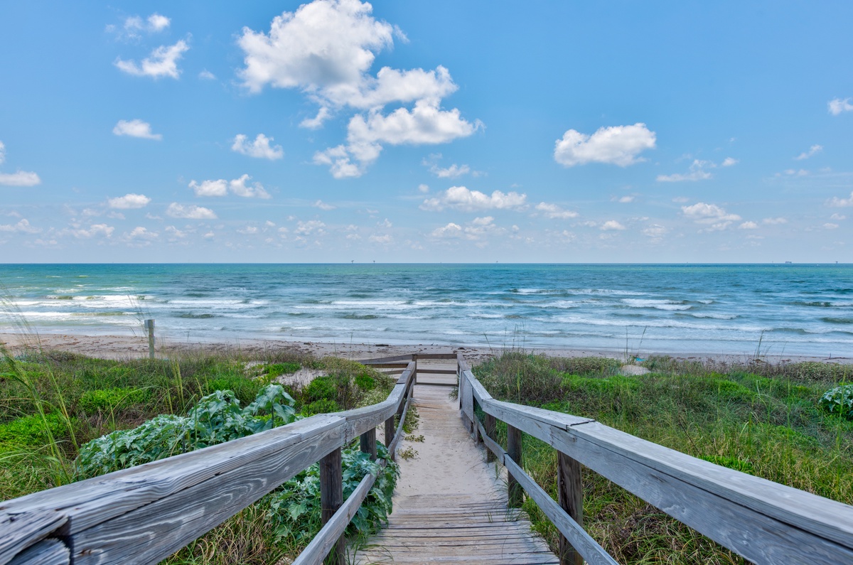 Wooden boardwalk leads through coastal dunes to pristine sandy beach with rolling waves under brilliant blue sky.
