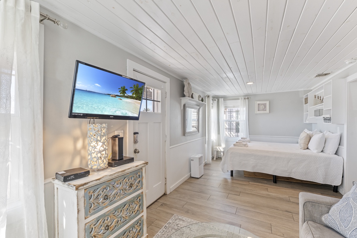 Unwind in your serene bedroom where white linens meet coastal charm, complete with a flat-screen TV and beautiful natural light streaming through elegant windows.