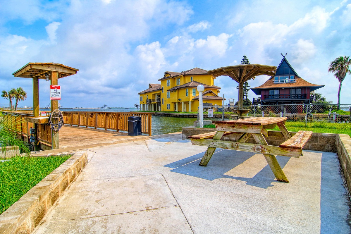 Waterfront picnic area with wooden tables and covered pier, featuring colorful coastal homes across the water.