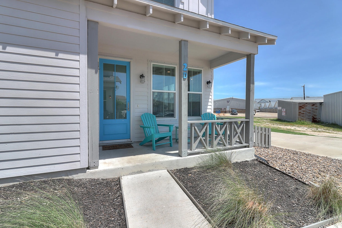 Modern coastal home with welcoming blue door and covered porch featuring turquoise chair in peaceful neighborhood setting.
