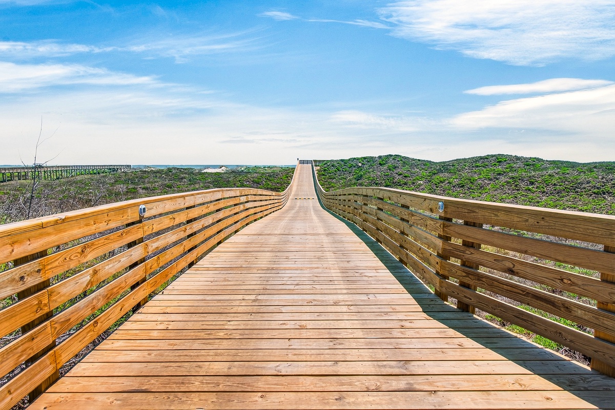 Wooden boardwalk extends toward the coastline through natural dunes and native vegetation under a bright blue sky.