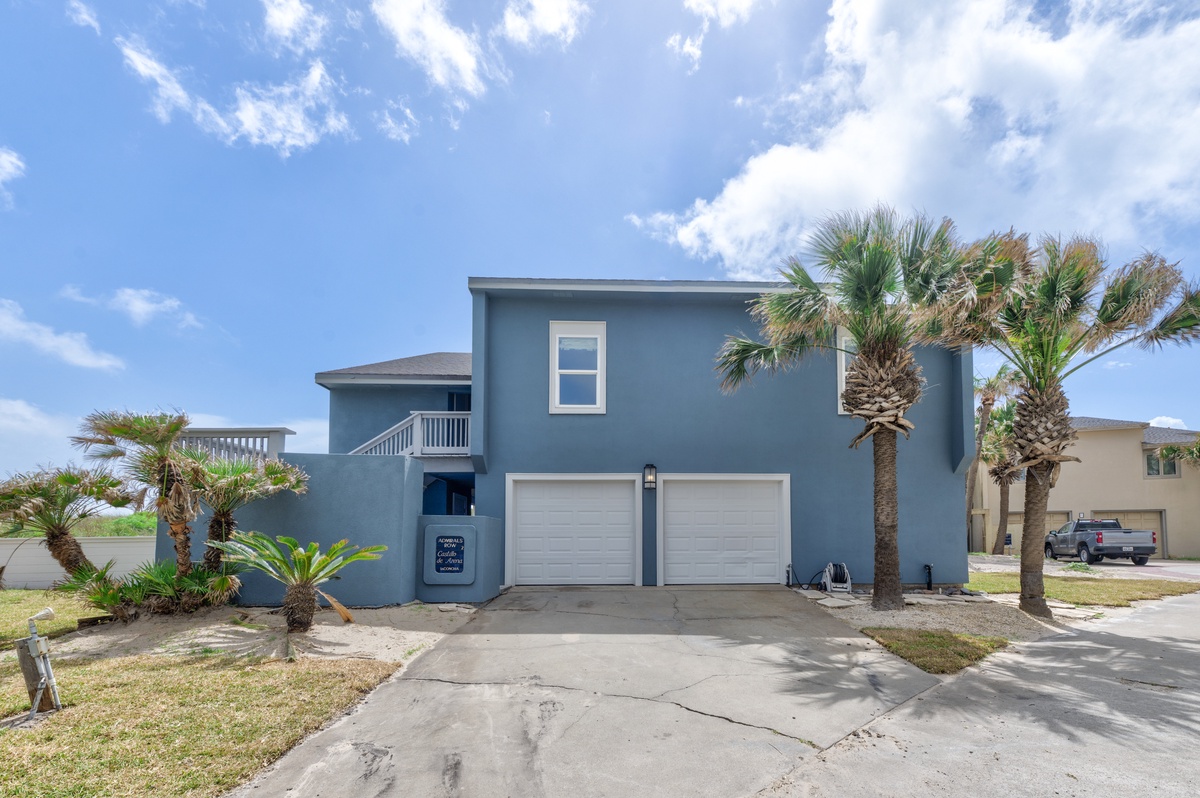 Two-story blue coastal home with palm trees and garage parking in a tropical residential neighborhood.