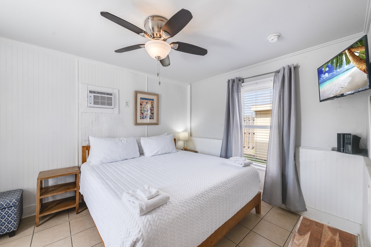 Your cozy bedroom features crisp white linens, gentle ceiling fan, and tropical TV for relaxing evenings after beach adventures.