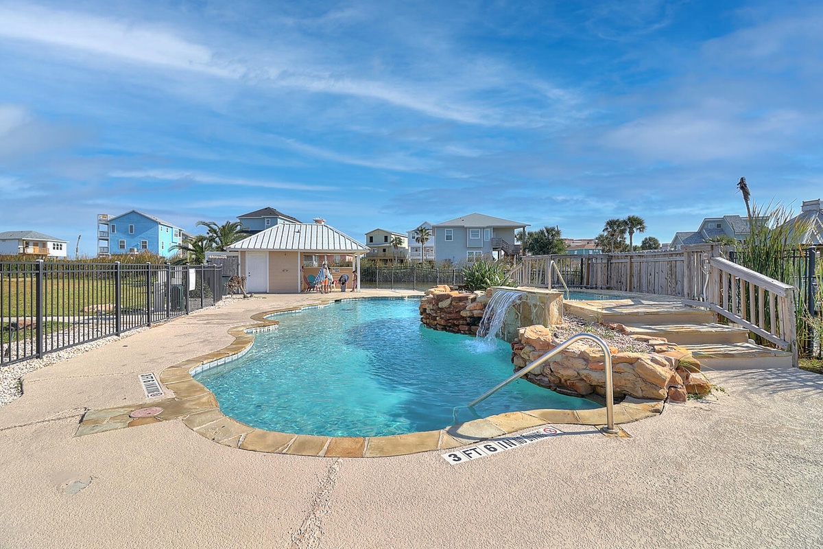 Sparkling community pool with waterfall feature surrounded by coastal homes under sunny blue skies.