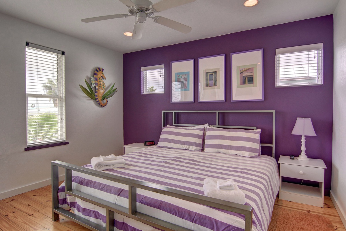Unwind in your vibrant bedroom featuring a bold purple accent wall, comfortable queen bed with crisp linens, and tropical touches that create a cheerful retreat.