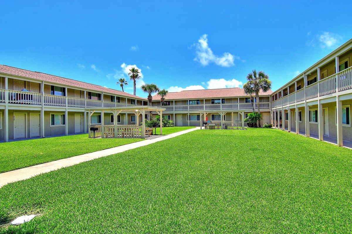 Property buildings form a welcoming courtyard with lush green lawn and tropical palm trees under bright blue skies.