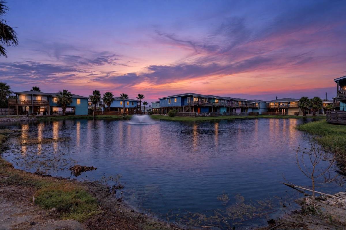 A peaceful pond fronted by modern vacation rentals captures the soft pastels of dusk, with palm trees and warm window lights adding to the coastal retreat atmosphere.