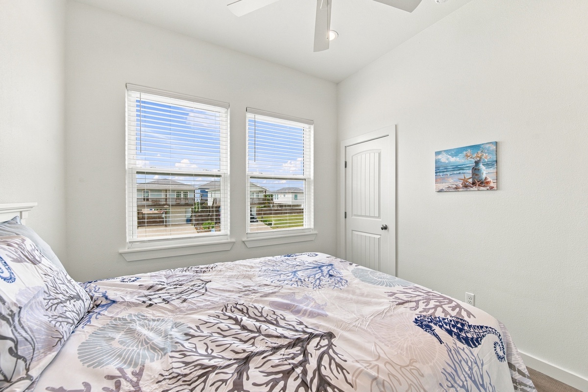 Drift into restful sleep in your primary bedroom with a queen bed, where sunlight filters through crisp white blinds and gentle beach artwork sets a serene, vacation mood.