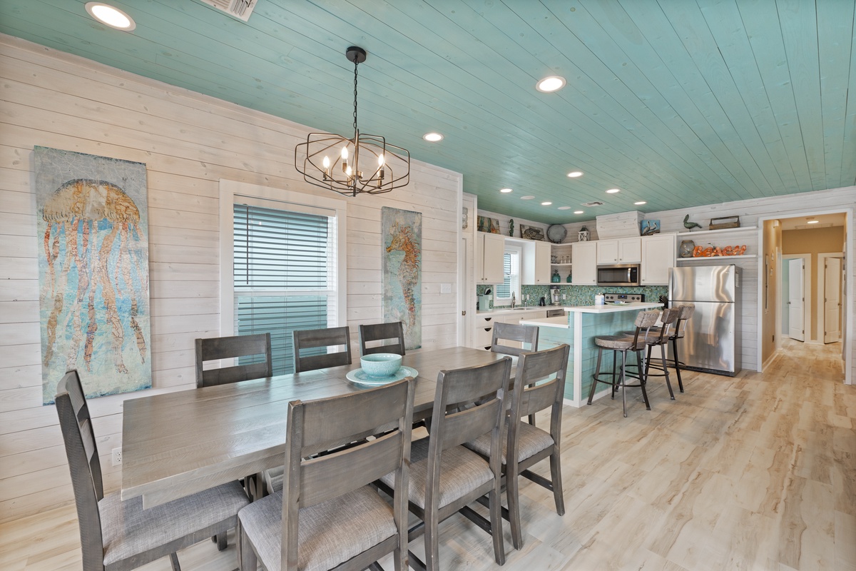 Step into this bright coastal dining space with stylish shiplap walls, a welcoming table for eight, and a charming open kitchen with bar seating. The soft turquoise ceiling and coastal touches make every meal feel like a beachside escape.