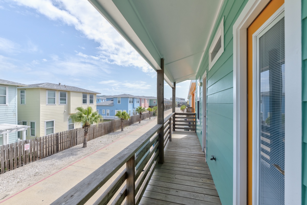 A covered wooden deck overlooks a quiet coastal neighborhood, offering glimpses of palm trees and colorful beach homes just beyond the railing.